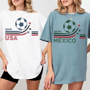 May include: Two short-sleeve t-shirts, one white and one dusty blue, each featuring a soccer ball graphic, curved stripes, stars, and text. The white shirt displays 'SOCCER USA' in red and blue, while the blue shirt displays 'SOCCER M&Eacute;XICO' in green and red.
