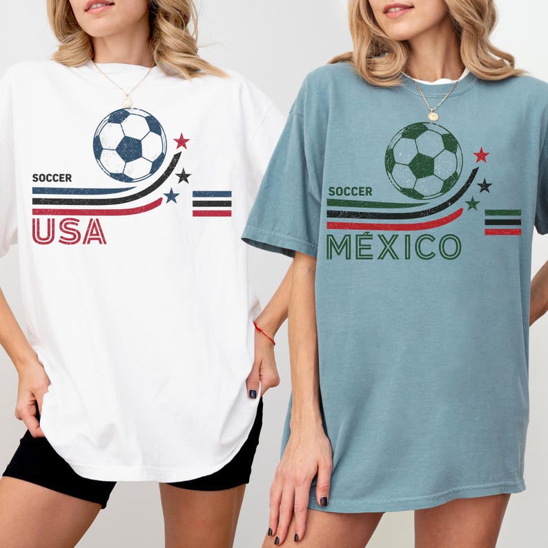 May include: Two short-sleeve t-shirts, one white and one dusty blue, each featuring a soccer ball graphic, curved stripes, stars, and text. The white shirt displays 'SOCCER USA' in red and blue, while the blue shirt displays 'SOCCER M&Eacute;XICO' in green and red.