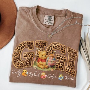 Könnte beinhalten: Braunes Comfort Colors T-Shirt mit dem Wort "GIGI" im Leopardenmuster, mit Winnie Puuh und einem Honigtopf. Darunter die Namen Emily, Robert, Sofia und Karen mit Bienengrafiken. Eine silberne Halskette ist über dem Shirt drapiert.