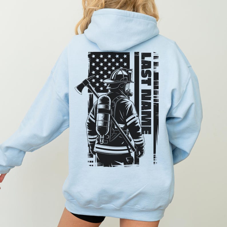 Op de afbeelding: Lichtblauwe hoodie met een brandweerman-afbeelding op de achterkant. Het ontwerp toont een brandweerman met een bijl en luchttank, tegen een Amerikaanse vlag en de tekst "LAST NAME".