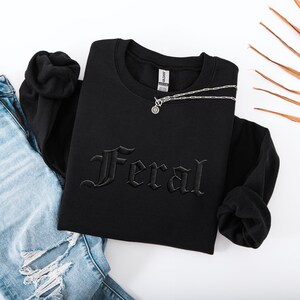 May include: Black crewneck sweatshirt with the word "Feral" embroidered in gothic lettering. A silver necklace with a circular pendant rests on the sweatshirt. The sweatshirt is paired with blue jeans.