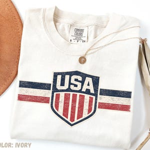 May include: An ivory short-sleeve t-shirt featuring a distressed graphic of a shield with the text USA above red and white vertical stripes. The design includes horizontal navy blue and red stripes extending from the shield. A gold pendant necklace rests on the shirt.