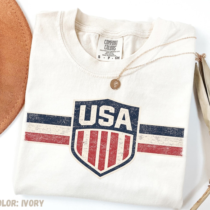 May include: An ivory short-sleeve t-shirt featuring a distressed graphic of a shield with the text USA above red and white vertical stripes. The design includes horizontal navy blue and red stripes extending from the shield. A gold pendant necklace rests on the shirt.