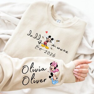 Pode incluir: Camisola bege com a inscri&ccedil;&atilde;o preta "Daddy Mouse Est. 2026" e um desenho do Mickey Mouse. A manga apresenta os nomes "Olivia" e "Oliver" com ilustra&ccedil;&otilde;es da Minnie e do Mickey Mouse. Inclui um cora&ccedil;&atilde;o e abelhas.