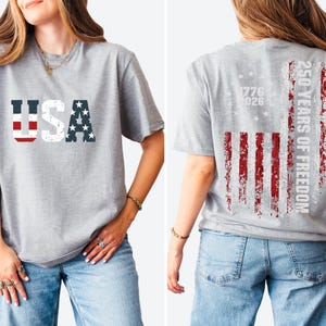May include: A heather gray t-shirt featuring the text USA on the front in a distressed American flag pattern. The back displays a vertical distressed American flag graphic with the text 250 YEARS OF FREEDOM and the years 1776 and 2026.