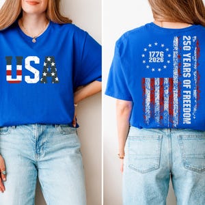 May include: A royal blue t-shirt featuring a distressed USA graphic on the front with red stripes and white stars. The back displays a distressed American flag design with a circle of stars, the text 1776 2026, and 250 YEARS OF FREEDOM in white.