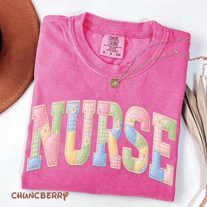 Puede incluir: Una camiseta rosa intenso con la palabra "NURSE" aplicada en la parte delantera con letras de patchwork. Las letras son de colores pastel con detalles de margaritas. Un collar dorado con un colgante descansa sobre la camiseta. La marca "CHUNCBERRY" está en la parte inferior.