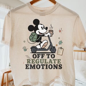 May include: An ivory-colored t-shirt featuring a graphic of Mickey Mouse riding a scooter while holding a coffee cup. The design includes a green backpack, a clipboard labeled PLAN, a puzzle piece, a handprint, and the text OFF TO REGULATE EMOTIONS.