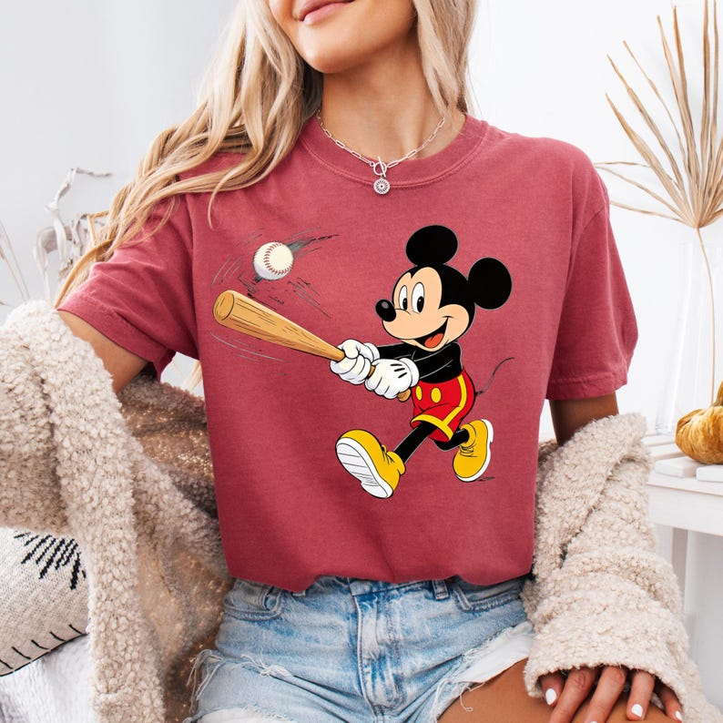 May include: A maroon t-shirt with a cartoon image of Mickey Mouse playing baseball. Mickey Mouse is depicted swinging a bat at a baseball. He wears yellow shoes, red shorts, white gloves, and a black shirt. The baseball is white.