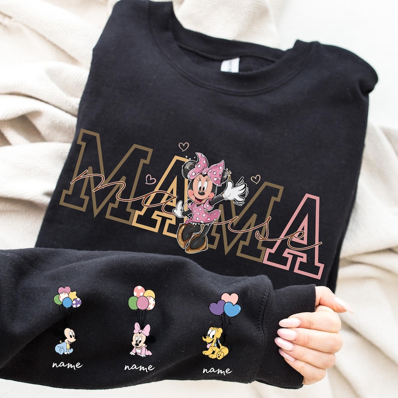 May include: Black sweatshirt featuring the word "MAMA" in gold and pink, with a Minnie Mouse graphic. The sleeve displays cartoon characters holding balloons, with space for names. The design includes hearts.