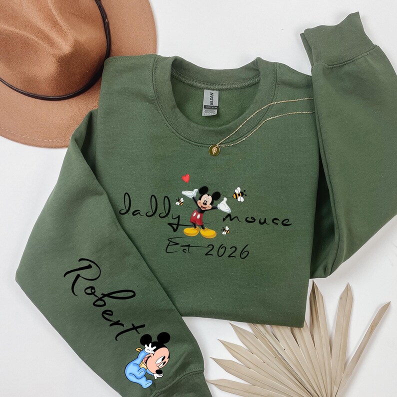 Pode incluir: Um moletom verde-oliva com as palavras "Daddy Mouse" e "Est 2026" em escrita preta. Um gr&aacute;fico do Mickey Mouse est&aacute; no centro, com um beb&ecirc; Mickey Mouse na manga. Um colar de ouro est&aacute; drapeado sobre o moletom.