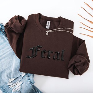 May include: A brown sweatshirt with the word "Feral" embroidered in black gothic lettering. A silver necklace with a pendant is draped across the sweatshirt. The sweatshirt is folded next to a pair of blue jeans.