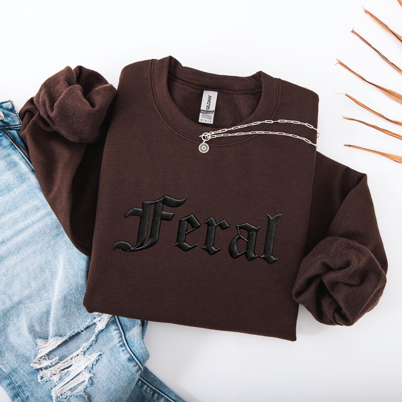May include: A brown sweatshirt with the word "Feral" embroidered in black gothic lettering. A silver necklace with a pendant is draped across the sweatshirt. The sweatshirt is folded next to a pair of blue jeans.