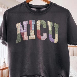 May include: A charcoal gray short-sleeve t-shirt featuring the letters NICU in a large, collegiate-style font. Each letter is constructed from quilted fabric patches in various patterns, including floral, gingham, and solid colors like green, purple, and pink.