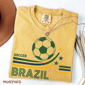 May include: A mustard yellow t-shirt featuring a graphic of a soccer ball above the text SOCCER BRAZIL. The design includes green, blue, and yellow curved lines with three small stars. The inner neck label reads COMFORT COLORS S P CH.