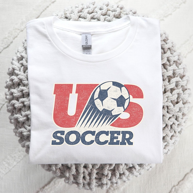 May include: A white short-sleeved t-shirt featuring a graphic design with the red letters US and the word SOCCER in blue. A black and white soccer ball is positioned over the letters with motion lines trailing behind it, all in a distressed style.