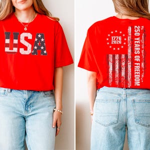 May include: A bright red short-sleeve t-shirt. The front features the text USA in a distressed flag pattern. The back displays a circular arrangement of stars above the years 1776 and 2026, with a vertical flag stripe and the text 250 YEARS OF FREEDOM.