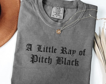 May include: A gray Comfort Colors t-shirt with the text "A Little Ray of Pitch Black" in black lettering. A gold-colored necklace with a pendant is draped across the shirt. The shirt is folded and displayed with a brown hat.