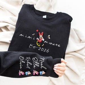 Pode incluir: Camisola preta com um design da Minnie Mouse e o texto "mimi mouse Est 2026". A manga exibe nomes e gr&aacute;ficos da Minnie Mouse. O design inclui detalhes em vermelho, branco e amarelo.