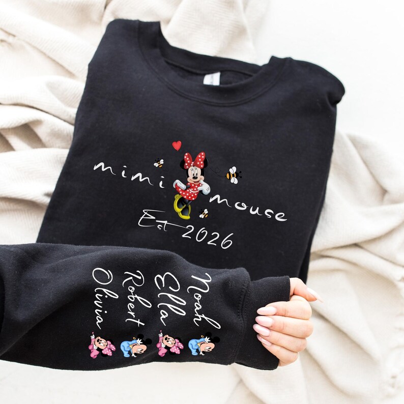 Pode incluir: Camisola preta com um design da Minnie Mouse e o texto "mimi mouse Est 2026". A manga exibe nomes e gr&aacute;ficos da Minnie Mouse. O design inclui detalhes em vermelho, branco e amarelo.