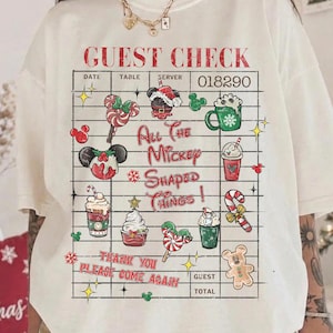 Pode incluir: Uma t-shirt cor de creme com um gráfico de uma conta de restaurante com o texto GUEST CHECK, All The Mickey SHAPED THINGS!, e THANK YOU PLEASE COME AGAIN. O design inclui várias ilustrações com tema natalício com ícones do Mickey.