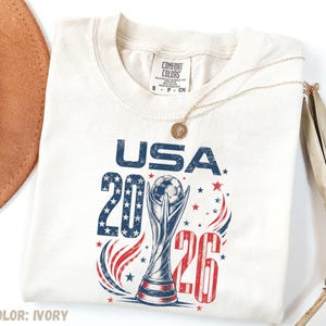 K&ouml;nnte beinhalten: Ein elfenbeinfarbenes T-Shirt mit einer Grafik mit dem Text USA 2026, einer Fu&szlig;ball-Troph&auml;en-Illustration sowie roten und blauen Sternen. Das T-Shirt hat ein Comfort Colors-Markenetikett am Kragen und eine goldfarbene Halskette liegt auf dem Stoff.