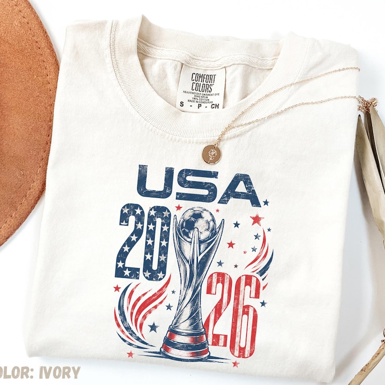 K&ouml;nnte beinhalten: Ein elfenbeinfarbenes T-Shirt mit einer Grafik mit dem Text USA 2026, einer Fu&szlig;ball-Troph&auml;en-Illustration sowie roten und blauen Sternen. Das T-Shirt hat ein Comfort Colors-Markenetikett am Kragen und eine goldfarbene Halskette liegt auf dem Stoff.