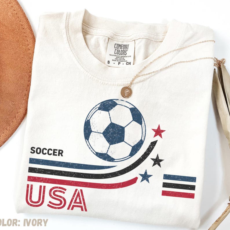 May include: An ivory t-shirt featuring a graphic of a soccer ball, three curved stripes in blue, black, and red, three stars, and the text "SOCCER USA". The shirt has a Comfort Colors brand label at the neck and a small gold pendant necklace.