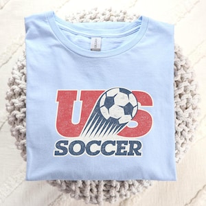 May include: A light blue t-shirt featuring a graphic with the red letters US, a black and white soccer ball with motion lines, and the word SOCCER in dark blue text below. The design has a distressed, vintage-inspired texture throughout.