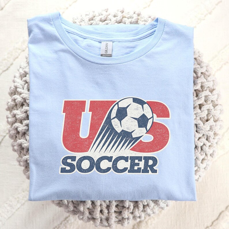 May include: A light blue t-shirt featuring a graphic with the red letters US, a black and white soccer ball with motion lines, and the word SOCCER in dark blue text below. The design has a distressed, vintage-inspired texture throughout.