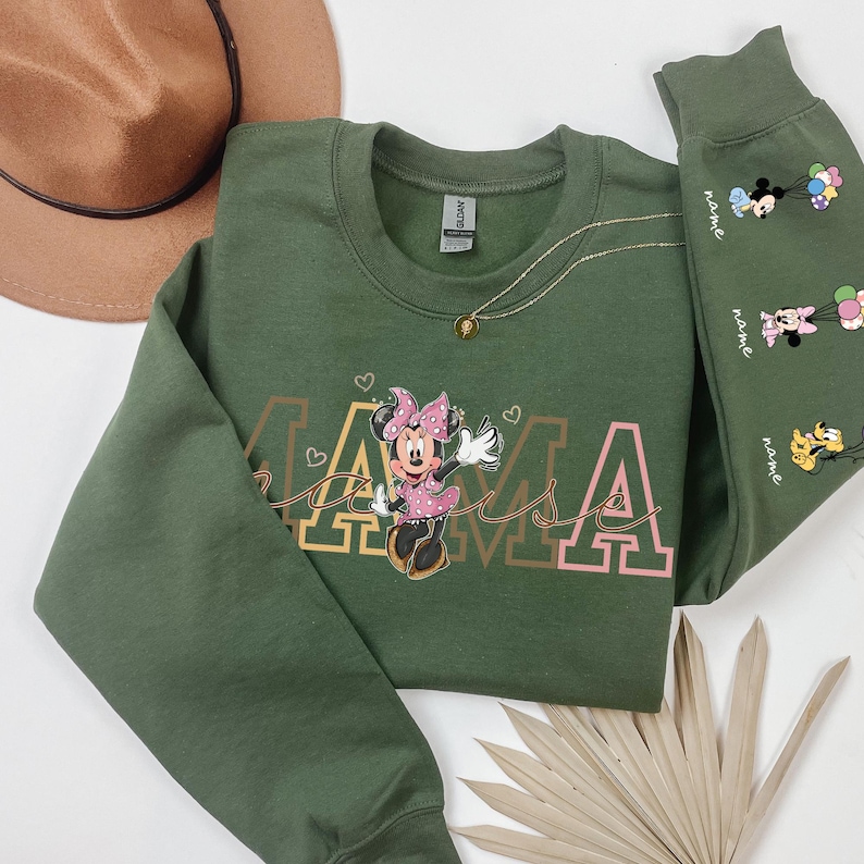 May include: Green sweatshirt with the word "MAMA" in a gold and pink font, featuring a Minnie Mouse graphic. The sleeve has cartoon characters with balloons. A gold necklace and a brown hat are also visible.
