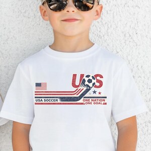 May include: A white short-sleeved t-shirt featuring a graphic design with the text USA SOCCER ONE NATION ONE GOAL. The design includes a soccer ball, a small American flag, and red, white, and blue stripes with a distressed, vintage-style texture.