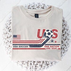 May include: A beige crew neck t-shirt featuring a graphic with the text USA, USA SOCCER, and ONE NATION ONE GOAL. The design includes a soccer ball, a United States flag, and red, white, and blue horizontal stripes with a distressed vintage texture.