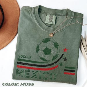 May include: A moss green t-shirt featuring a distressed graphic of a soccer ball above the text SOCCER M&Eacute;XICO. The design includes curved stripes in green, black, and red, three small stars, and a horizontal bar pattern in the same colors.