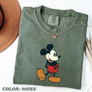 May include: A moss green t-shirt featuring a graphic of Mickey Mouse in his classic pose. The shirt has a crew neck and short sleeves. A gold-colored necklace with a pendant rests on the shirt. The text "COLOR: MOSS" is at the bottom.