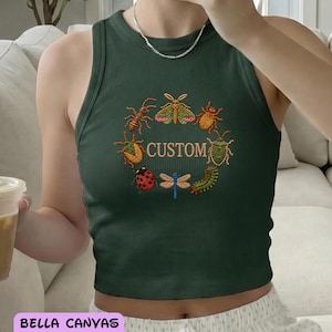 May include: A dark green ribbed racerback tank top featuring an embroidered circular design of various colorful insects surrounding the word CUSTOM in the center. The text BELLA CANVAS RACER TANK is printed in a pink box at the bottom left.