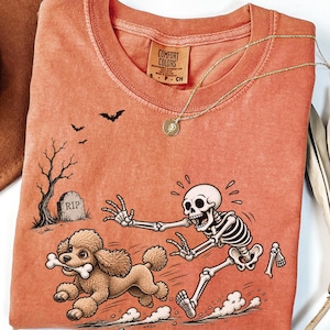 May include: A terracotta-coloured t-shirt featuring a graphic of a skeleton chasing a brown poodle that is carrying a bone in its mouth. The scene includes a tombstone marked RIP, a bare tree, and bats. The shirt has a Comfort Colors brand label.