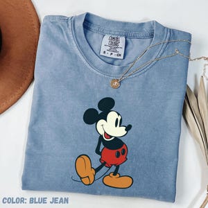 May include: A blue jean colored t-shirt featuring a graphic of Mickey Mouse. The shirt has a crew neck and short sleeves. A gold necklace with a pendant is draped across the shirt. The text "COLOR: BLUE JEAN" is at the bottom.