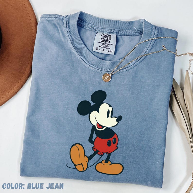 May include: A blue jean colored t-shirt featuring a graphic of Mickey Mouse. The shirt has a crew neck and short sleeves. A gold necklace with a pendant is draped across the shirt. The text "COLOR: BLUE JEAN" is at the bottom.