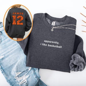 Puede incluir: Sudadera gris oscuro con el texto blanco "apparently, i like basketball". La parte trasera de la sudadera tiene el nombre "JAMES" y el número "12" en naranja, con un diseño de baloncesto. Un collar plateado está sobre la sudadera.