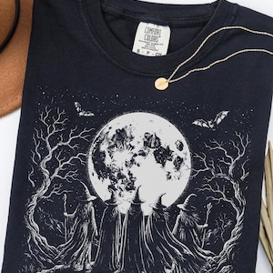 Può includere: Una t-shirt nera a girocollo con una stampa grafica bianca di cinque figure in mantello e cappello a punta davanti a una grande luna piena. La scena include alberi spogli e pipistrelli in volo. L'etichetta interna riporta Comfort Colors.