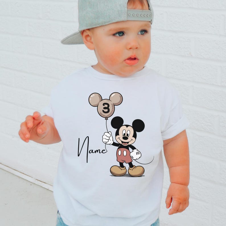 May include: White t-shirt with a Mickey Mouse design holding a balloon with the number 3. The balloon is beige, and the shirt has the name "Name" printed in cursive. The t-shirt is worn by a child.