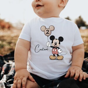 May include: White t-shirt featuring a Mickey Mouse graphic holding a balloon with the number 2. The balloon is beige and black. The word "Custom" is written in cursive below the balloon. The shirt is worn by a child.