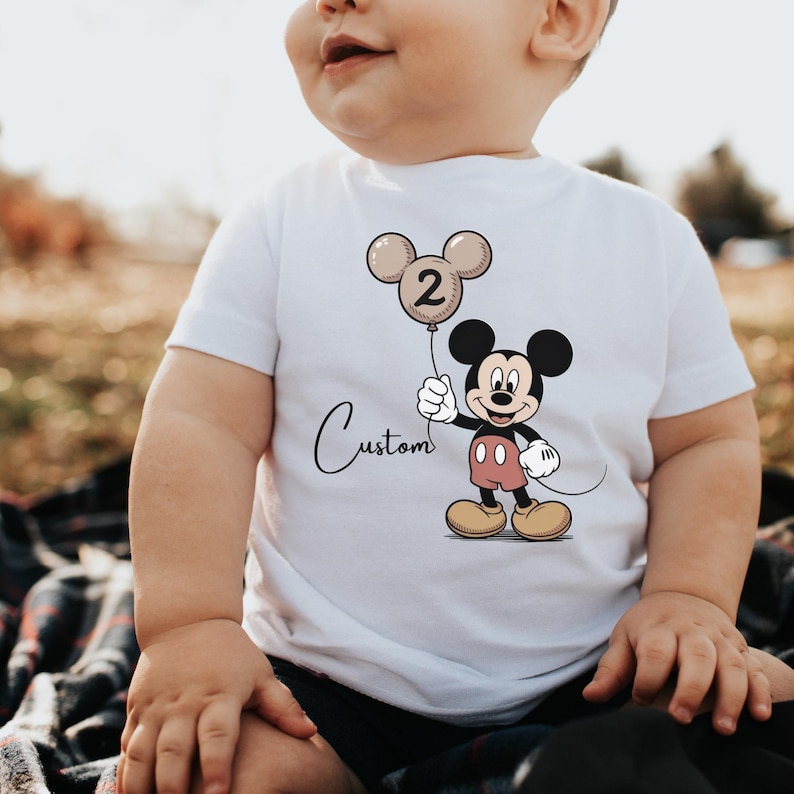 May include: White t-shirt featuring a Mickey Mouse graphic holding a balloon with the number 2. The balloon is beige and black. The word "Custom" is written in cursive below the balloon. The shirt is worn by a child.