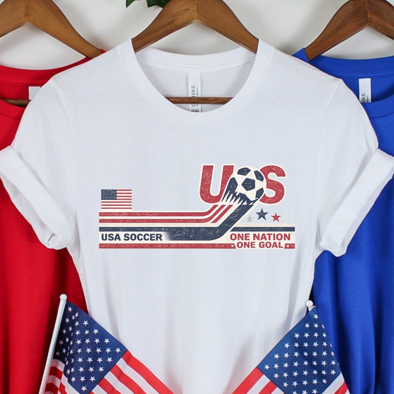 May include: A white short-sleeve t-shirt featuring a graphic with the text USA SOCCER ONE NATION ONE GOAL. The design includes a soccer ball, a small American flag, and red, white, and blue stripes with a distressed, vintage-style texture.