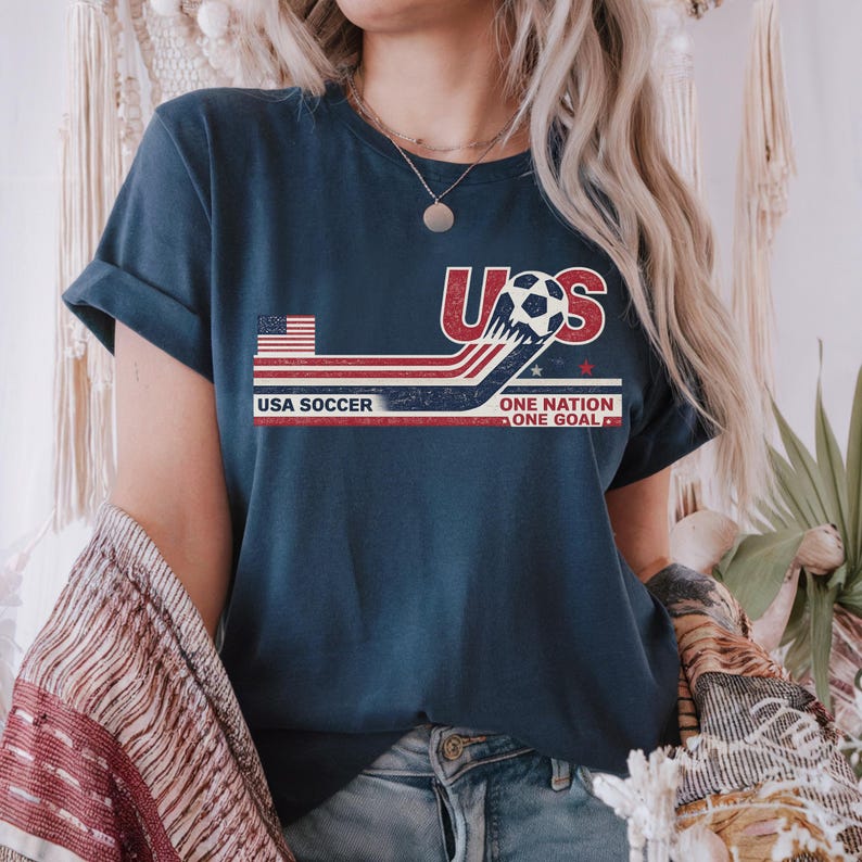 May include: A dark blue t-shirt featuring a graphic with the text USA, USA SOCCER, and ONE NATION ONE GOAL. The design includes a small American flag, a soccer ball, and red, white, and blue horizontal stripes with a distressed, vintage texture.