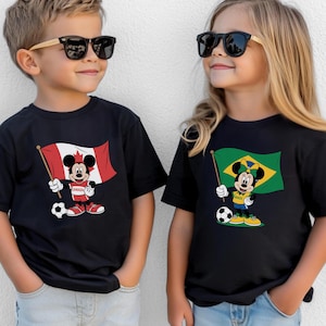 May include: Two black t-shirts featuring cartoon characters. The left shirt displays Mickey Mouse holding a Canadian flag and a soccer ball. The right shirt displays Minnie Mouse holding a Brazilian flag and a soccer ball. Both shirts have short sleeves.