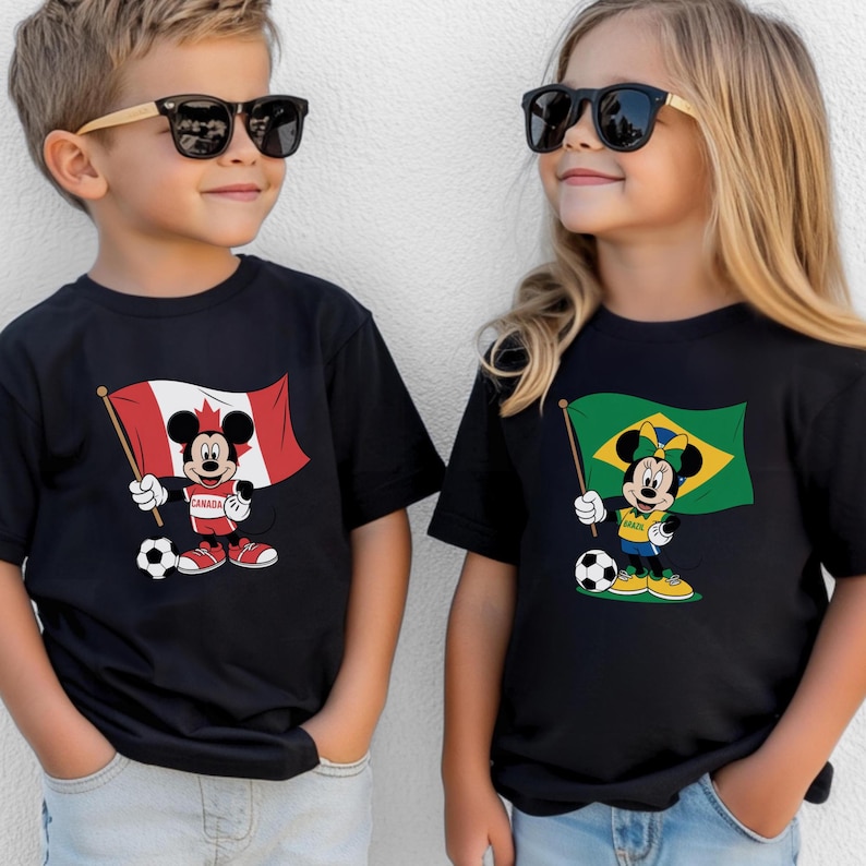 May include: Two black t-shirts featuring cartoon characters. The left shirt displays Mickey Mouse holding a Canadian flag and a soccer ball. The right shirt displays Minnie Mouse holding a Brazilian flag and a soccer ball. Both shirts have short sleeves.