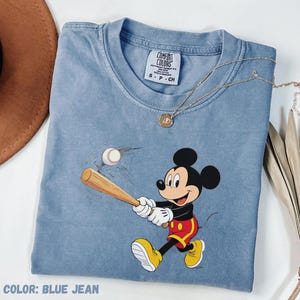 May include: Blue jean-coloured t-shirt featuring a graphic of Mickey Mouse swinging a baseball bat. The shirt has a crew neck and short sleeves. A gold necklace with a pendant is draped across the shirt. The text "COLOR: BLUE JEAN" is at the bottom.