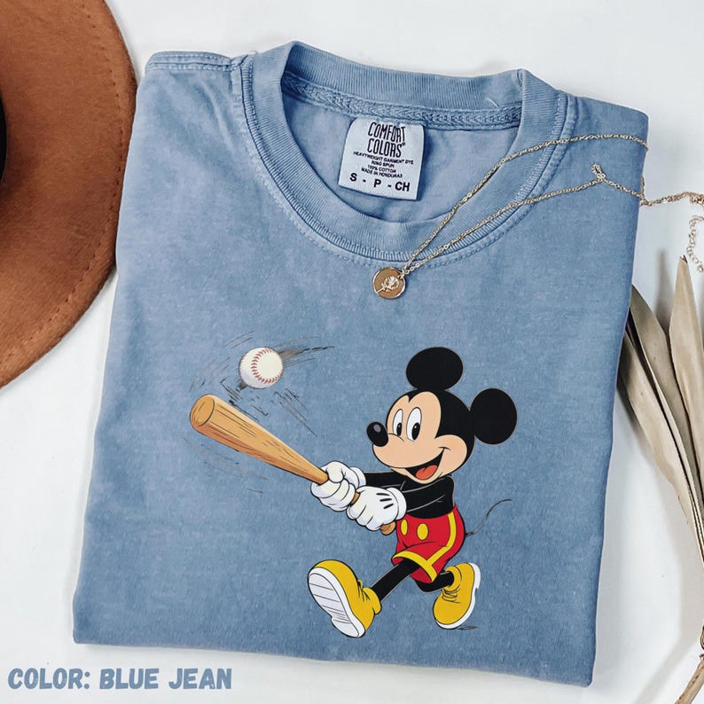 May include: Blue jean-coloured t-shirt featuring a graphic of Mickey Mouse swinging a baseball bat. The shirt has a crew neck and short sleeves. A gold necklace with a pendant is draped across the shirt. The text "COLOR: BLUE JEAN" is at the bottom.